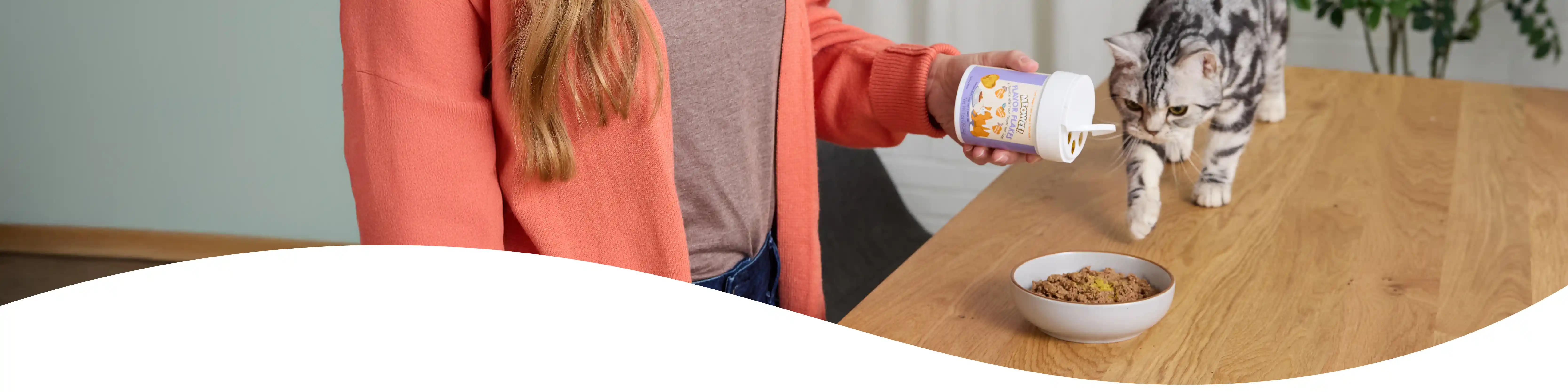 Woman shaking Meowee cat topper flakes onto cat food, cat walking toward bowl